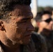 A close-up photograph of a military service member with sand covering his face and hair, suggesting recent physical training or a beach/surf exercise. He is wearing camouflage uniform and appears focused or fatigued. Other service members in camouflage are visible in the blurred background.