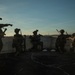 A group of military personnel in full tactical gear are positioned on a rooftop at dusk or dawn, appearing to conduct a tactical operation or training exercise. The soldiers are equipped with helmets, night vision devices, body armor, and weapons. One soldier on the left is pointing in a direction, suggesting coordination or command communication.