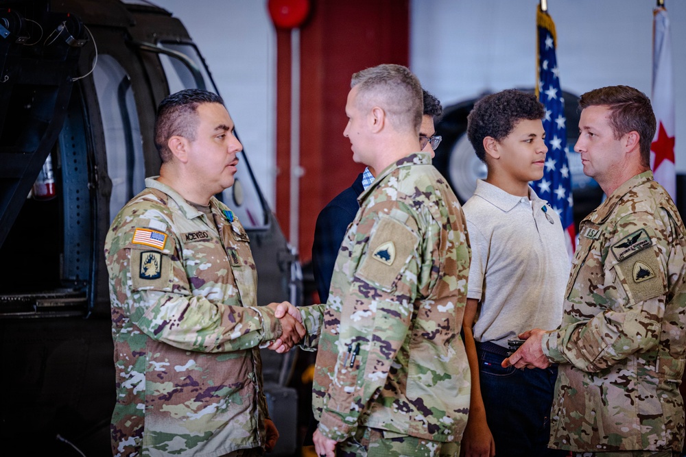 A military ceremony taking place inside what appears to be a hangar or military facility, where a soldier identified by name tape as 'ACEVEDO' is shaking hands with another uniformed soldier. Multiple individuals are present, including civilians, suggesting a formal recognition or award ceremony. An American flag and what appears to be a District of Columbia flag are visible in the background.