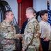 A military ceremony taking place inside what appears to be a hangar or military facility, where a soldier identified by name tape as 'ACEVEDO' is shaking hands with another uniformed soldier. Multiple individuals are present, including civilians, suggesting a formal recognition or award ceremony. An American flag and what appears to be a District of Columbia flag are visible in the background.
