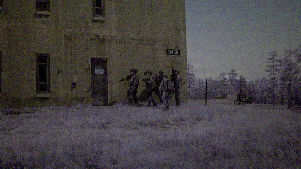An infrared or night-vision style photograph showing a group of armed, uniformed personnel moving along the exterior wall of a two-story brick building numbered '9450'. The scene appears to be a military or law enforcement training exercise conducted in a rural or semi-abandoned area. The distinctive purple-gray tonal rendering suggests the image was captured with infrared photography or a specialized camera filter.