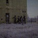 An infrared or night-vision style photograph showing a group of armed, uniformed personnel moving along the exterior wall of a two-story brick building numbered '9450'. The scene appears to be a military or law enforcement training exercise conducted in a rural or semi-abandoned area. The distinctive purple-gray tonal rendering suggests the image was captured with infrared photography or a specialized camera filter.