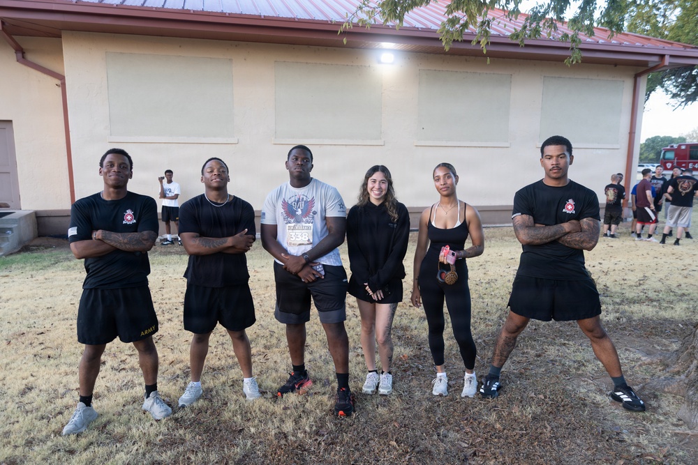 Six young adults pose together outdoors in front of a tan building with a red roof, appearing to be on a military installation or similar facility. The group consists of four males and two females, dressed in athletic/casual wear, with at least one person wearing Army-branded clothing. Several additional people are visible in the background near what appears to be a fire truck.