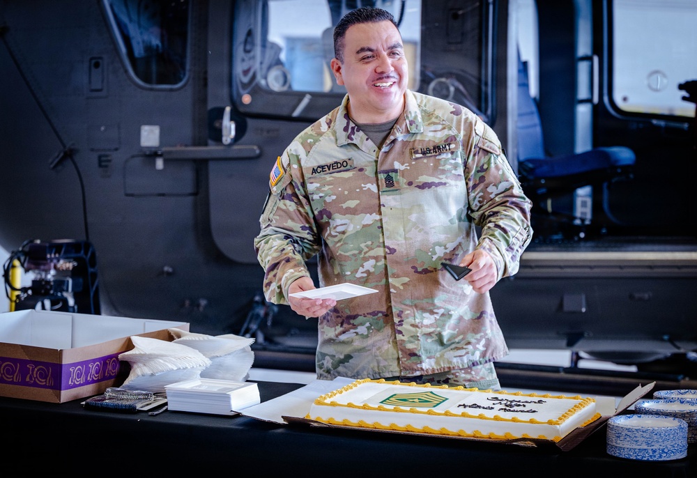 A U.S. Army soldier named Acevedo, wearing OCP (Operational Camouflage Pattern) uniform, stands smiling behind a table with a decorated sheet cake. The setting appears to be inside a military hangar or vehicle bay, suggesting a military celebration or promotion ceremony. A Sergeant Major rank insignia is visible on the cake, indicating this may be a promotion or retirement event.