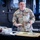 Sgt. Maj. Antonio Acevedo Promotion Ceremony A U.S. Army soldier named Acevedo, wearing OCP (Operational Camouflage Pattern) uniform, stands smiling behind a table with a decorated sheet cake. The setting appears to be inside a military hangar or vehicle bay, suggesting a military celebration or promotion ceremony. A Sergeant Major rank insignia is visible on the cake, indicating this may be a promotion or retirement event.