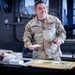 A U.S. Army soldier named Acevedo, wearing OCP (Operational Camouflage Pattern) uniform, stands smiling behind a table with a decorated sheet cake. The setting appears to be inside a military hangar or vehicle bay, suggesting a military celebration or promotion ceremony. A Sergeant Major rank insignia is visible on the cake, indicating this may be a promotion or retirement event.