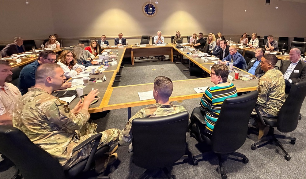 A formal conference or briefing session is taking place in a large meeting room, with approximately 25-30 attendees seated around a U-shaped table arrangement. Several participants are wearing military camouflage uniforms (OCP/MultiCam pattern), while others are in civilian business attire. A military emblem or seal is visible on the wall at the far end of the room.