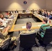 A formal conference or briefing session is taking place in a large meeting room, with approximately 25-30 attendees seated around a U-shaped table arrangement. Several participants are wearing military camouflage uniforms (OCP/MultiCam pattern), while others are in civilian business attire. A military emblem or seal is visible on the wall at the far end of the room.