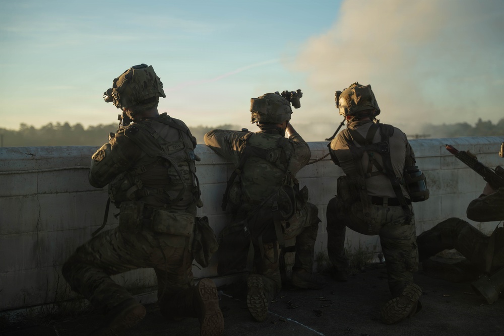 Three military personnel in full combat gear are positioned behind a low concrete wall or barrier, appearing to observe or assess a situation ahead. They are photographed from behind in a low-light or dusk environment, with visible smoke or haze in the background. The scene suggests a tactical operation, training exercise, or active engagement scenario.