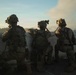 Three military personnel in full combat gear are positioned behind a low concrete wall or barrier, appearing to observe or assess a situation ahead. They are photographed from behind in a low-light or dusk environment, with visible smoke or haze in the background. The scene suggests a tactical operation, training exercise, or active engagement scenario.