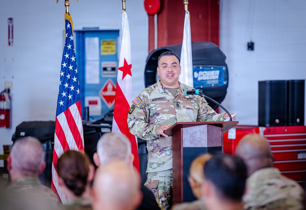 A U.S. Army soldier named Acevedo, wearing OCP (Operational Camouflage Pattern) uniform, is delivering a speech at a podium in what appears to be a military maintenance bay or motor pool facility. The American flag and the District of Columbia flag are visible behind the speaker. An audience of uniformed military personnel is seated in the foreground listening to the address.