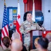 A U.S. Army soldier named Acevedo, wearing OCP (Operational Camouflage Pattern) uniform, is delivering a speech at a podium in what appears to be a military maintenance bay or motor pool facility. The American flag and the District of Columbia flag are visible behind the speaker. An audience of uniformed military personnel is seated in the foreground listening to the address.