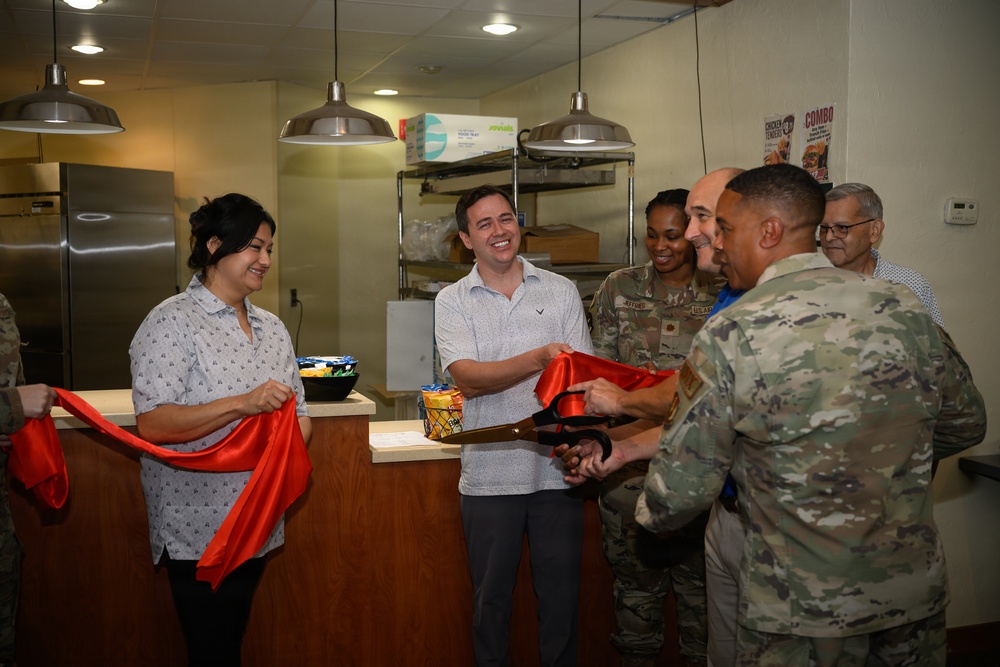 A ribbon-cutting ceremony is taking place inside what appears to be a military base food service facility or snack bar. Several individuals, including military personnel in camouflage uniforms and civilians, are gathered together using large ceremonial scissors to cut a red ribbon. The atmosphere is celebratory, suggesting the opening or reopening of a dining or food service establishment.