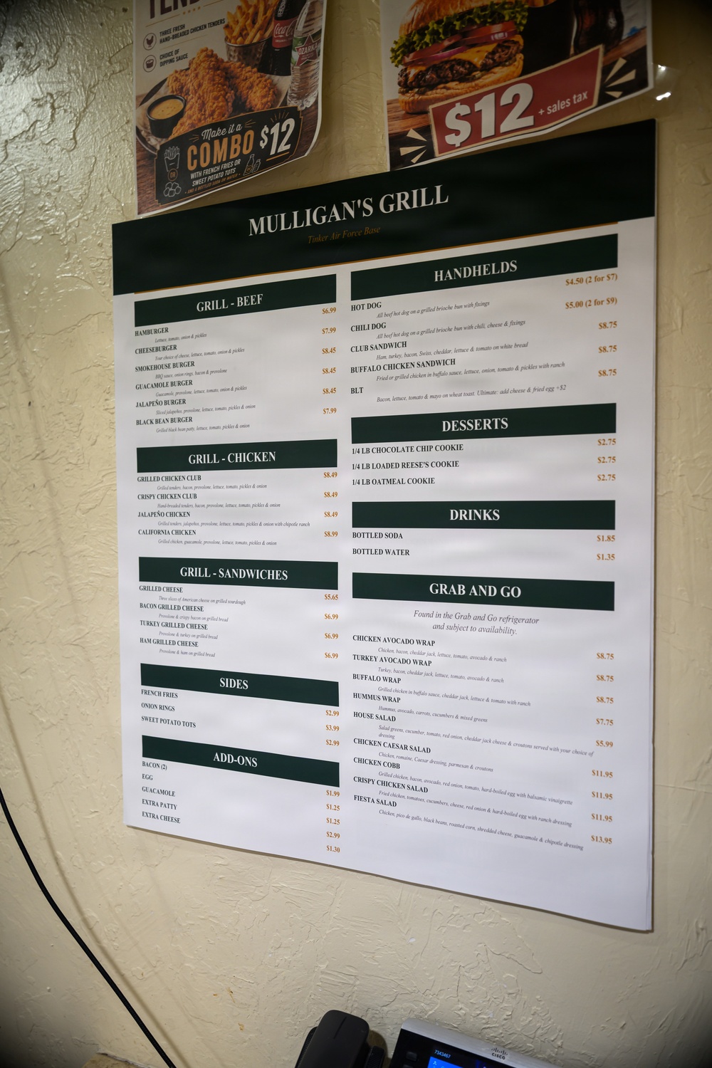 A wall-mounted menu for Mulligan's Grill located at Tinker Air Force Base is displayed on a white textured wall. The menu is organized into multiple sections covering beef burgers, chicken, sandwiches, sides, add-ons, handhelds, desserts, drinks, and grab-and-go items. Above the main menu are two promotional signs advertising combo deals at $12.