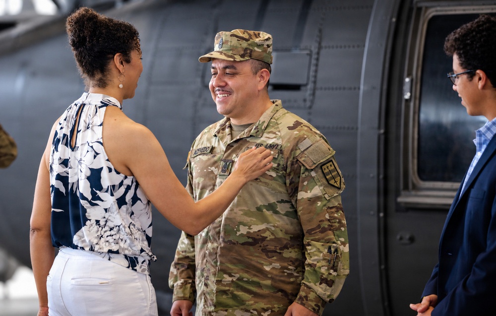 A U.S. Army soldier in OCP (Operational Camouflage Pattern) uniform is smiling warmly while a woman places her hand on his chest, appearing to pin or adjust something on his uniform. The setting appears to be a military hangar or airfield, with a large military aircraft visible in the background. A younger male in civilian clothes observes from the right side of the frame.