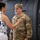 Sgt. Maj. Antonio Acevedo Promotion Ceremony A U.S. Army soldier in OCP (Operational Camouflage Pattern) uniform is smiling warmly while a woman places her hand on his chest, appearing to pin or adjust something on his uniform. The setting appears to be a military hangar or airfield, with a large military aircraft visible in the background. A younger male in civilian clothes observes from the right side of the frame.