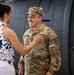 A U.S. Army soldier in OCP (Operational Camouflage Pattern) uniform is smiling warmly while a woman places her hand on his chest, appearing to pin or adjust something on his uniform. The setting appears to be a military hangar or airfield, with a large military aircraft visible in the background. A younger male in civilian clothes observes from the right side of the frame.