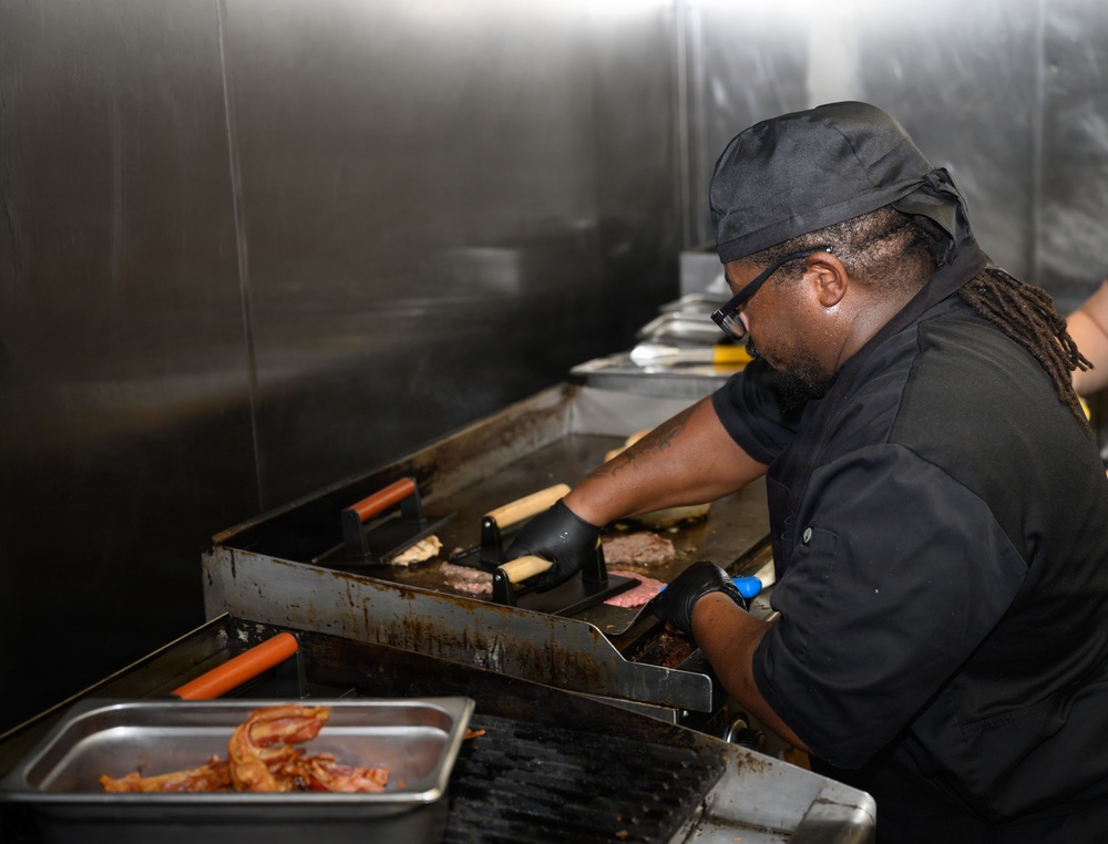 A cook wearing all-black uniform, black gloves, glasses, and a black head covering is working at a commercial flat-top griddle. They are actively cooking multiple items including burger patties, hot dogs, and what appears to be buns. The setting is a professional kitchen with stainless steel surfaces and a metal prep tray containing cooked bacon in the foreground.
