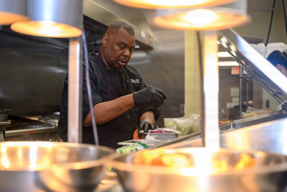 A professional chef dressed in a dark uniform and black gloves is preparing food at a commercial kitchen station. He is working under heat lamps, handling fresh ingredients including what appears to be vegetables and greens. The setting appears to be a restaurant or institutional kitchen with stainless steel equipment.