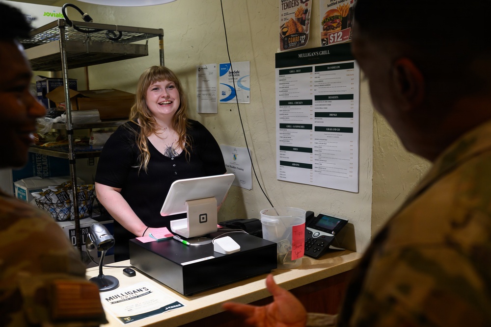 A smiling female cashier stands behind a counter at Mulligan's Grill, interacting with two individuals wearing military camouflage uniforms. The setting appears to be a small food service establishment, likely located on a military base. A Square point-of-sale system and cash drawer are visible on the counter.