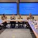 A formal military and civilian meeting is taking place in a conference room, identified as 'Mission Partner Conversations - Morning Round' involving OC-ALC, 72 ABW, 448 SCMW &amp; Software units. Participants include both uniformed military personnel in camouflage (OCP) uniforms and civilians seated around a U-shaped conference table arrangement. Two large blue presentation screens display the meeting title at the front of the room.