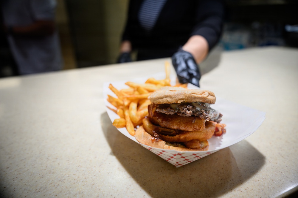 A food service worker wearing black nitrile gloves is presenting a large burger with onion rings and melted cheese, served alongside french fries in a red-checkered paper food tray. The meal is being placed on a light-colored counter surface. The burger appears to be a substantial double patty with multiple toppings.