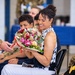 A woman in a black and white floral sleeveless top is seated at what appears to be a military ceremony, smiling as she receives a bouquet of mixed flowers from a soldier in camouflage uniform. Several other individuals, including children and military personnel, are seated nearby. The setting appears to be an indoor military event with blue-draped tables visible in the background.