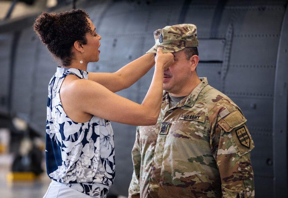 A woman in a floral dress is placing or adjusting a military cap on a U.S. Army soldier's head, likely during a promotion or pinning ceremony. The soldier is wearing OCP (Operational Camouflage Pattern) uniform with U.S. Army tape and a unit patch visible on the right shoulder. The setting appears to be a military hangar or aircraft facility.