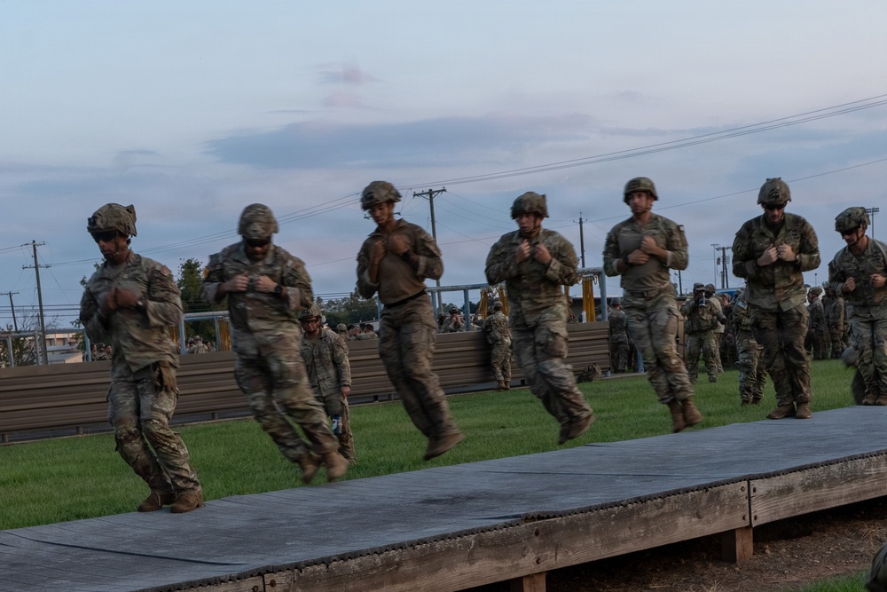A group of military personnel in camouflage uniforms and helmets are performing synchronized physical training exercises on a wooden platform at dusk. The soldiers appear to be executing a coordinated drill, possibly a combatives or physical readiness training exercise. A larger formation of soldiers is visible in the background observing or waiting their turn.