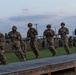 A group of military personnel in camouflage uniforms and helmets are performing synchronized physical training exercises on a wooden platform at dusk. The soldiers appear to be executing a coordinated drill, possibly a combatives or physical readiness training exercise. A larger formation of soldiers is visible in the background observing or waiting their turn.