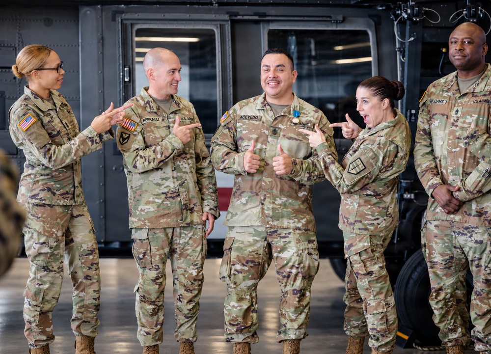 Five U.S. Army soldiers in OCP (Operational Camouflage Pattern) uniforms are gathered in what appears to be a military hangar, celebrating a fellow soldier named Acevedo who is wearing a medal. The group is in a jovial, celebratory mood with multiple soldiers pointing at Acevedo and giving thumbs up gestures. The setting appears to be an award ceremony or recognition event in an aviation hangar.