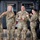 Sgt. Maj. Antonio Acevedo Promotion Ceremony Five U.S. Army soldiers in OCP (Operational Camouflage Pattern) uniforms are gathered in what appears to be a military hangar, celebrating a fellow soldier named Acevedo who is wearing a medal. The group is in a jovial, celebratory mood with multiple soldiers pointing at Acevedo and giving thumbs up gestures. The setting appears to be an award ceremony or recognition event in an aviation hangar.