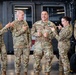 Five U.S. Army soldiers in OCP (Operational Camouflage Pattern) uniforms are gathered in what appears to be a military hangar, celebrating a fellow soldier named Acevedo who is wearing a medal. The group is in a jovial, celebratory mood with multiple soldiers pointing at Acevedo and giving thumbs up gestures. The setting appears to be an award ceremony or recognition event in an aviation hangar.