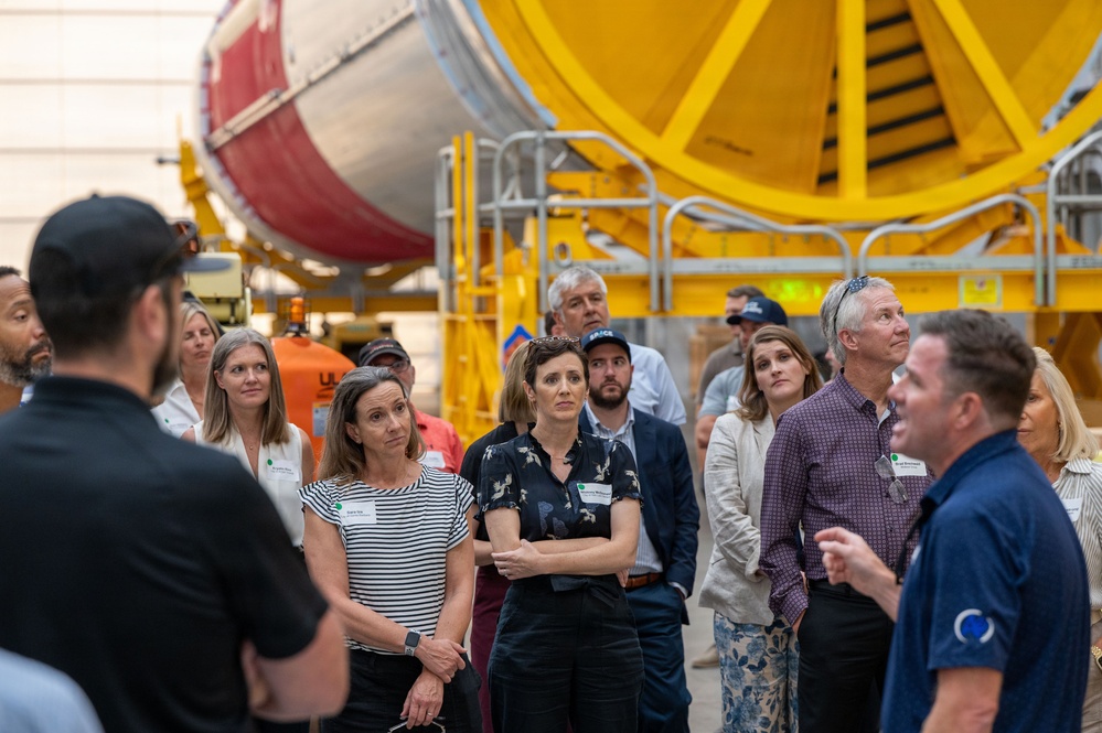 A group of visitors is receiving a guided tour inside what appears to be an aerospace or rocket manufacturing facility. A guide on the right is speaking to the attentive crowd, who are looking upward at large industrial equipment. In the background, a large red and white cylindrical rocket or fuel tank component is mounted on yellow industrial support structures.