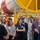 A group of visitors is receiving a guided tour inside what appears to be an aerospace or rocket manufacturing facility. A guide on the right is speaking to the attentive crowd, who are looking upward at large industrial equipment. In the background, a large red and white cylindrical rocket or fuel tank component is mounted on yellow industrial support structures.