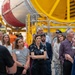 A group of visitors is receiving a guided tour inside what appears to be an aerospace or rocket manufacturing facility. A guide on the right is speaking to the attentive crowd, who are looking upward at large industrial equipment. In the background, a large red and white cylindrical rocket or fuel tank component is mounted on yellow industrial support structures.