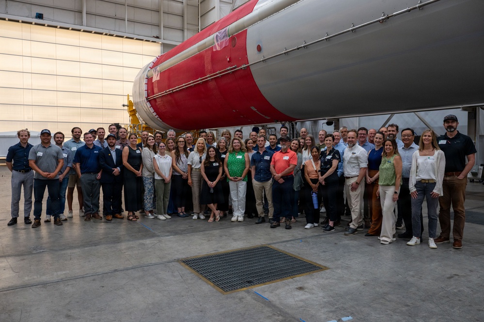A large group of approximately 40-50 people are posing for a group photograph inside what appears to be a rocket assembly or integration facility. They are standing in front of a large rocket or launch vehicle with a distinctive red and white color scheme. Many individuals are wearing name tags, suggesting this is an organized tour, company event, or professional gathering.
