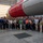 A large group of approximately 40-50 people are posing for a group photograph inside what appears to be a rocket assembly or integration facility. They are standing in front of a large rocket or launch vehicle with a distinctive red and white color scheme. Many individuals are wearing name tags, suggesting this is an organized tour, company event, or professional gathering.