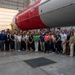A large group of approximately 40-50 people are posing for a group photograph inside what appears to be a rocket assembly or integration facility. They are standing in front of a large rocket or launch vehicle with a distinctive red and white color scheme. Many individuals are wearing name tags, suggesting this is an organized tour, company event, or professional gathering.