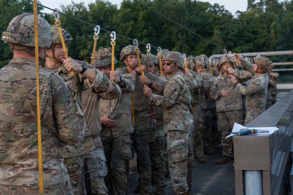 A line of U.S. Army soldiers in MultiCam/OCP camouflage uniforms and combat helmets are standing in formation on what appears to be a bridge or elevated platform, each holding yellow static line cables attached to an overhead wire. The scene appears to depict a pre-jump or airborne training exercise, with soldiers conducting equipment checks. A clipboard or paperwork is visible on a stand to the right, suggesting an official inspection or manifest process.