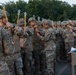 A line of U.S. Army soldiers in MultiCam/OCP camouflage uniforms and combat helmets are standing in formation on what appears to be a bridge or elevated platform, each holding yellow static line cables attached to an overhead wire. The scene appears to depict a pre-jump or airborne training exercise, with soldiers conducting equipment checks. A clipboard or paperwork is visible on a stand to the right, suggesting an official inspection or manifest process.
