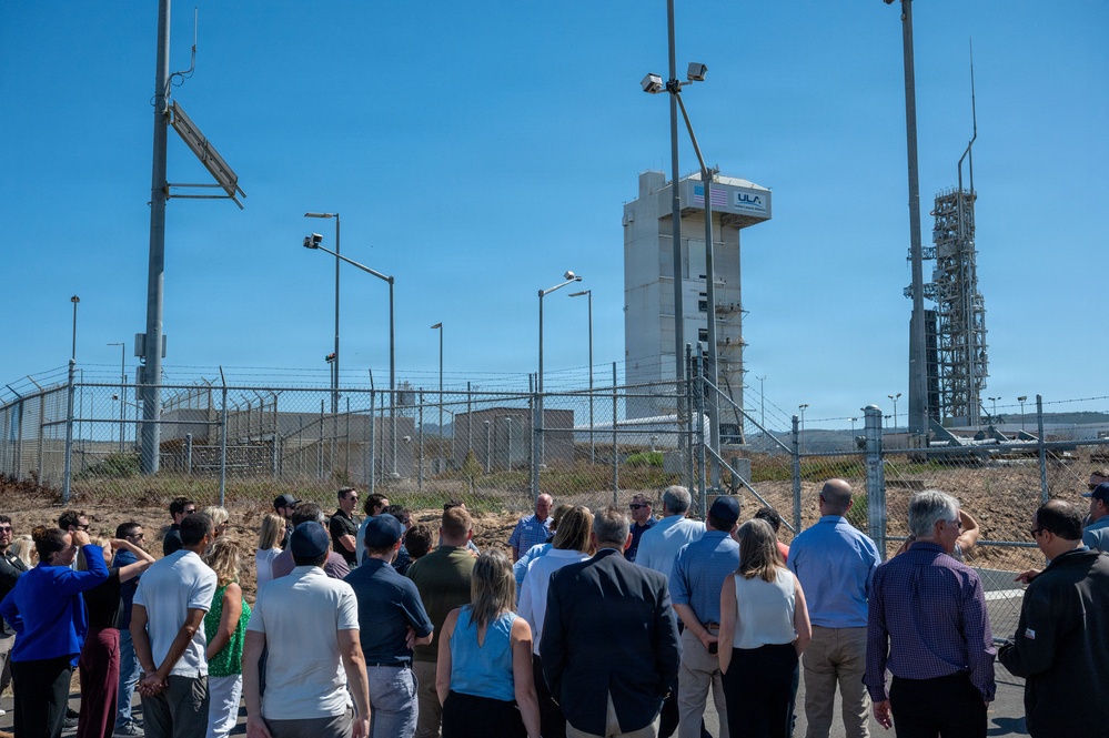 A group of approximately 30-40 people are gathered outdoors near a ULA (United Launch Alliance) launch facility, appearing to receive a briefing or tour. The scene is set at what appears to be Vandenberg Space Force Base in California, with a ULA launch tower and rocket infrastructure visible in the background behind a security fence. The group is facing one or two speakers positioned in the center of the crowd.