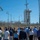 A group of approximately 30-40 people are gathered outdoors near a ULA (United Launch Alliance) launch facility, appearing to receive a briefing or tour. The scene is set at what appears to be Vandenberg Space Force Base in California, with a ULA launch tower and rocket infrastructure visible in the background behind a security fence. The group is facing one or two speakers positioned in the center of the crowd.