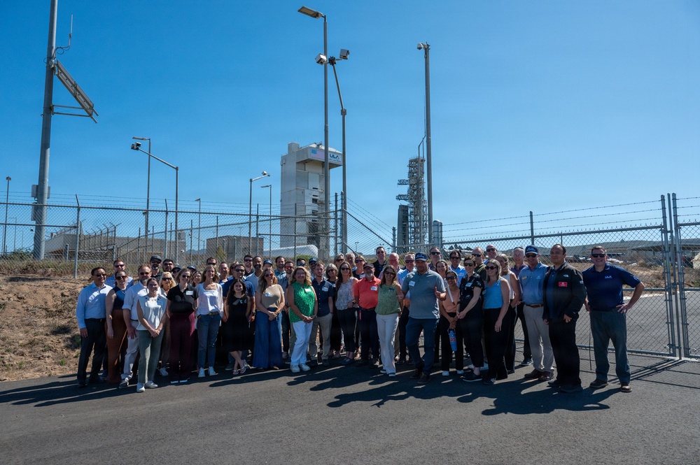 A large group of approximately 50-60 people pose for a group photo outdoors at what appears to be a ULA (United Launch Alliance) launch facility, likely Vandenberg Space Force Base in California. A white ULA-branded launch tower and rocket infrastructure are visible behind a chain-link security fence in the background. The group appears to be on an official tour or visit to the facility on a clear, sunny day.