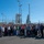 A large group of approximately 50-60 people pose for a group photo outdoors at what appears to be a ULA (United Launch Alliance) launch facility, likely Vandenberg Space Force Base in California. A white ULA-branded launch tower and rocket infrastructure are visible behind a chain-link security fence in the background. The group appears to be on an official tour or visit to the facility on a clear, sunny day.