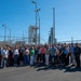 A large group of approximately 50-60 people pose for a group photo outdoors at what appears to be a ULA (United Launch Alliance) launch facility, likely Vandenberg Space Force Base in California. A white ULA-branded launch tower and rocket infrastructure are visible behind a chain-link security fence in the background. The group appears to be on an official tour or visit to the facility on a clear, sunny day.
