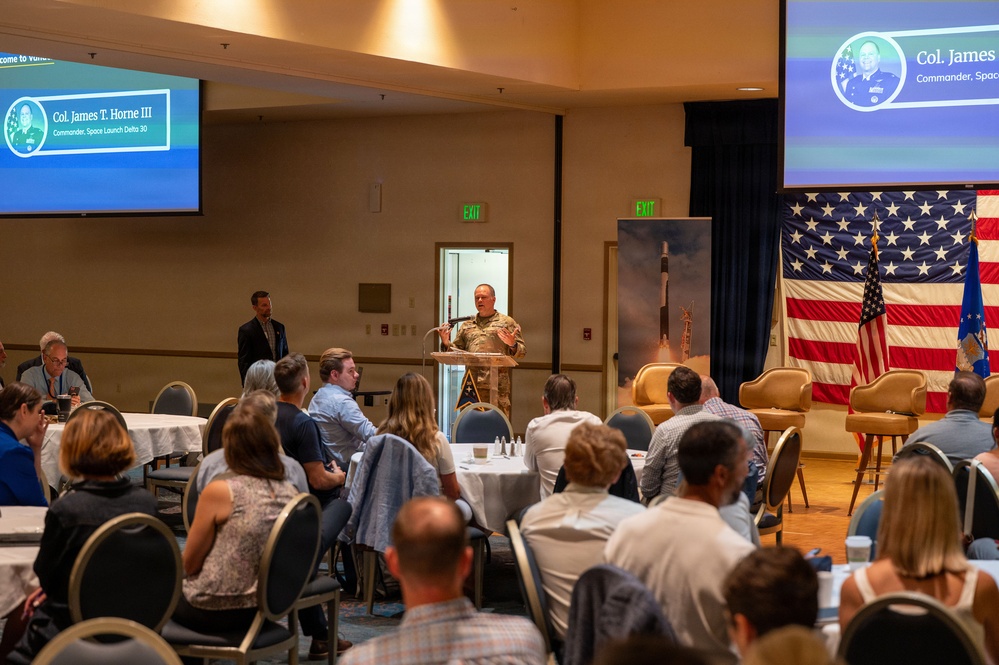 A military officer, identified by on-screen text as Col. James T. Horne III, Commander of Space Launch Delta 30, is delivering a presentation to a seated civilian audience in a banquet or conference hall. Two projection screens display his name, title, and photo. The setting includes American and Air Force flags, a rocket launch image backdrop, and round tables with attendees.