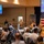 A military officer, identified by on-screen text as Col. James T. Horne III, Commander of Space Launch Delta 30, is delivering a presentation to a seated civilian audience in a banquet or conference hall. Two projection screens display his name, title, and photo. The setting includes American and Air Force flags, a rocket launch image backdrop, and round tables with attendees.