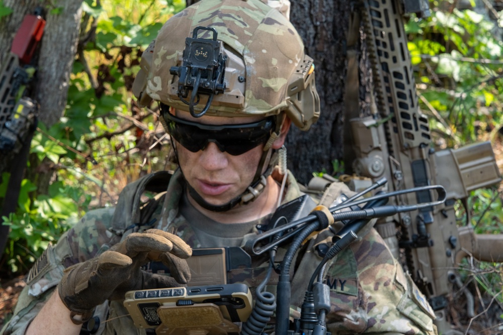 A U.S. Army soldier in full combat gear is operating a ruggedized tactical handheld device in a wooded outdoor environment. The soldier is wearing MultiCam camouflage, a ballistic helmet with mounted optics, and tactical gloves. Multiple communication cables and antennas are visible, suggesting active field communications or data operations.