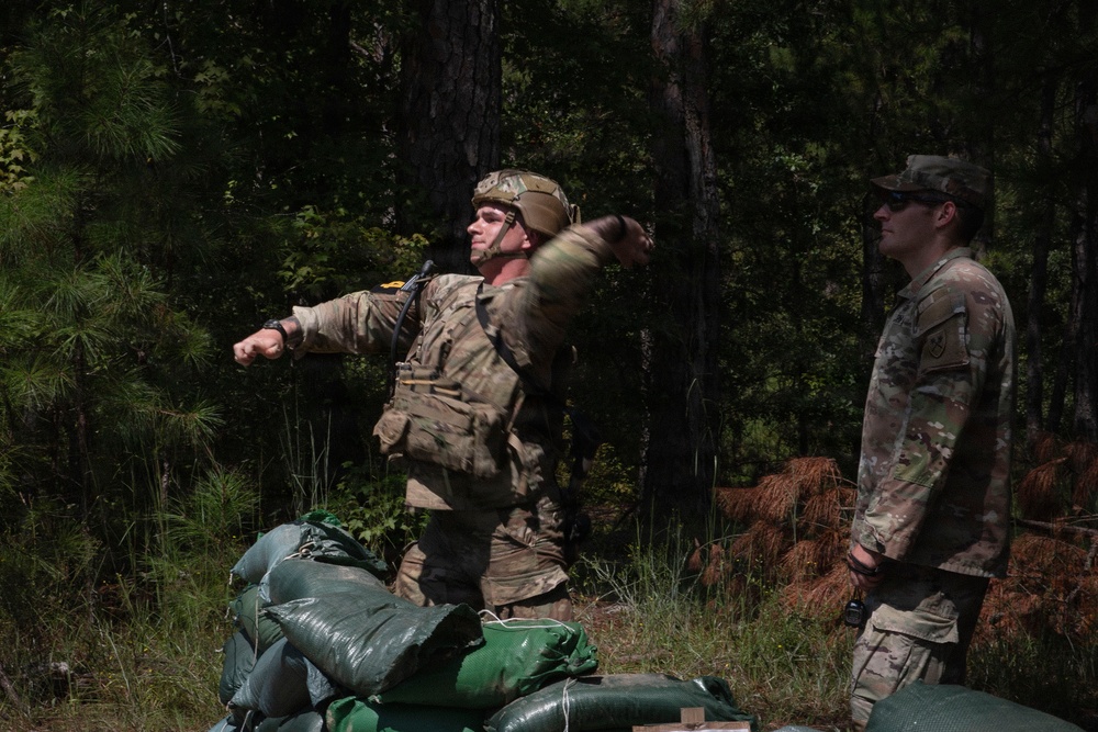 Two soldiers in US Army OCP (Operational Camouflage Pattern) uniforms are engaged in what appears to be a field training exercise in a wooded area. The soldier on the left, wearing a helmet and tactical vest, is in a throwing motion, likely practicing grenade throwing. A second soldier stands nearby observing, possibly acting as a safety observer or instructor.